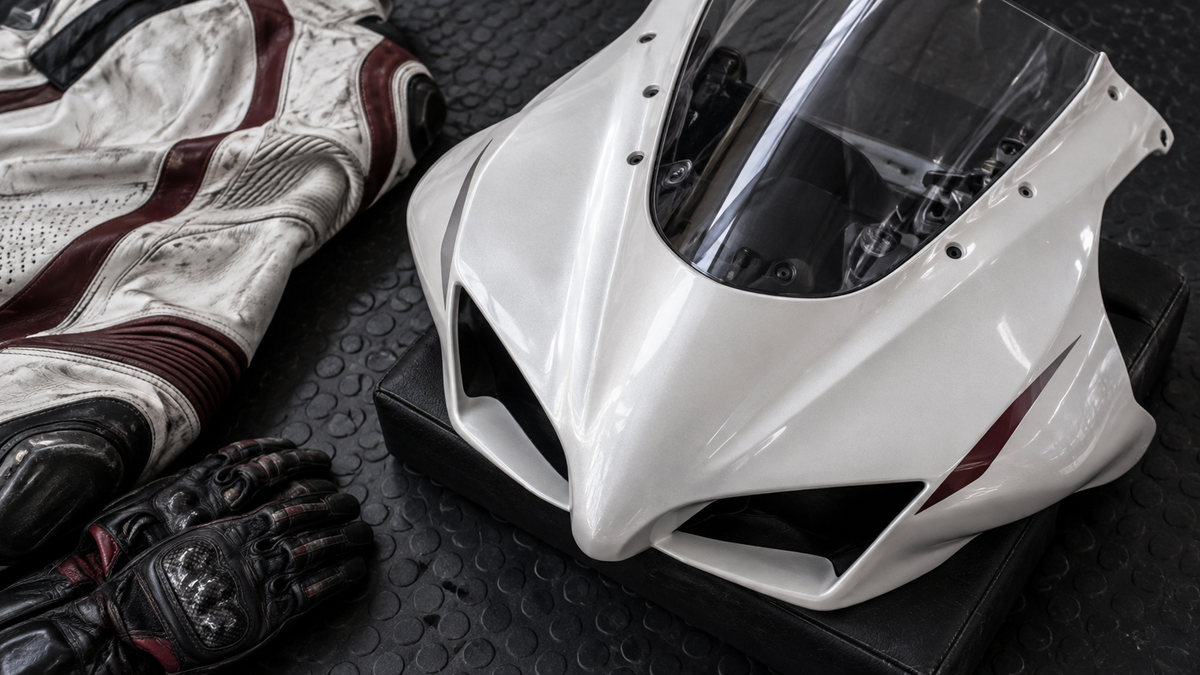 AI editorial concept: pearl-white nose fairing beside a folded racing suit and gloves; not official Aprilia hardware or a historical suit.